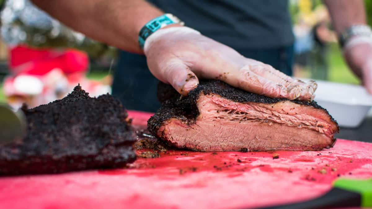 A gloved hand squashing cooked brisket onto a red cutting board