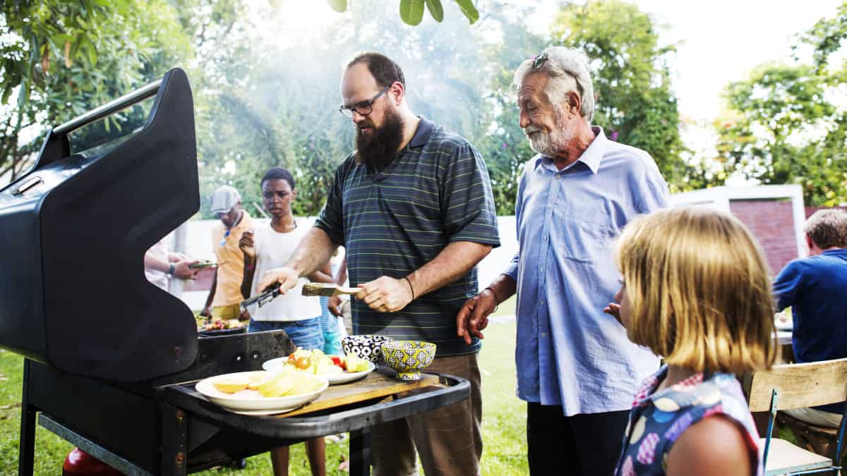 A family gathered around a BBQ while a man is cooking