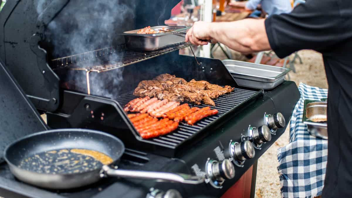 Sausages and meat on a gas grill, with a frying pan on the side burner