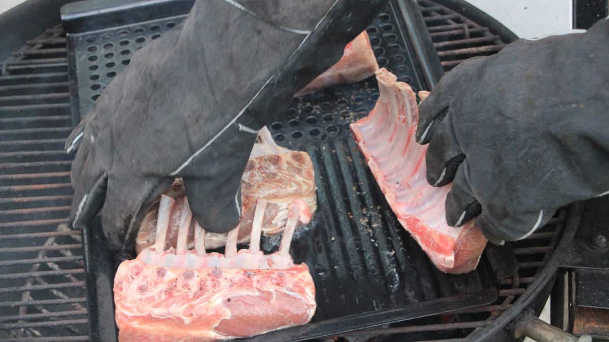 Close up of man wearing grilling bbq gloves while putting racks of lamb on the grill