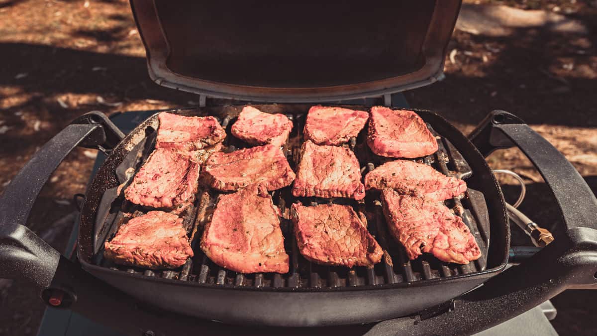 A black, portable gas grill with marinated beef cooking on the grates