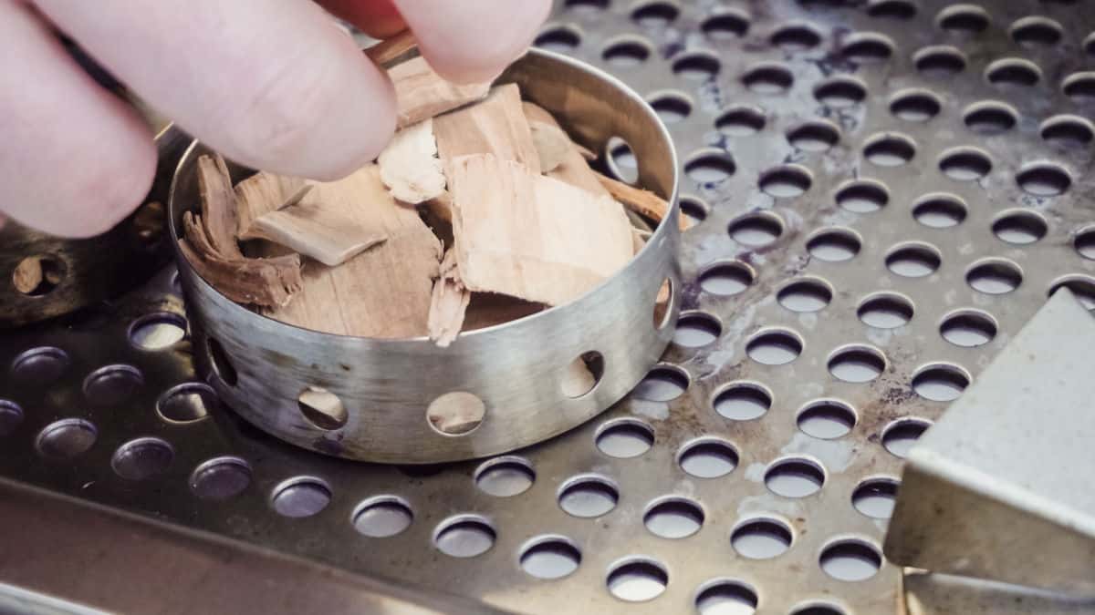 A round wood chip smoker box being filled on a gas grill