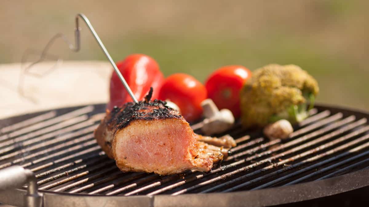 A remote Bluetooth thermometer probe stuck into some meat on a grill next to some vegetables