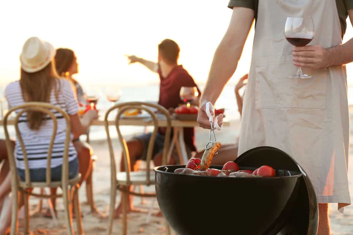 People sitting on the beach, one cooking on a portable charcoal grill