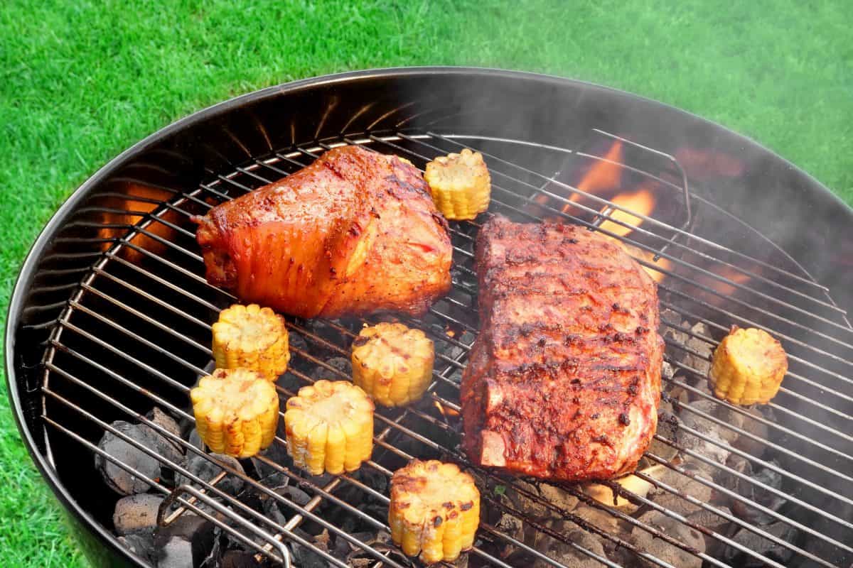 A charcoal kettle grill with 2 pieces of meat and some sweetcorn sections being grilled