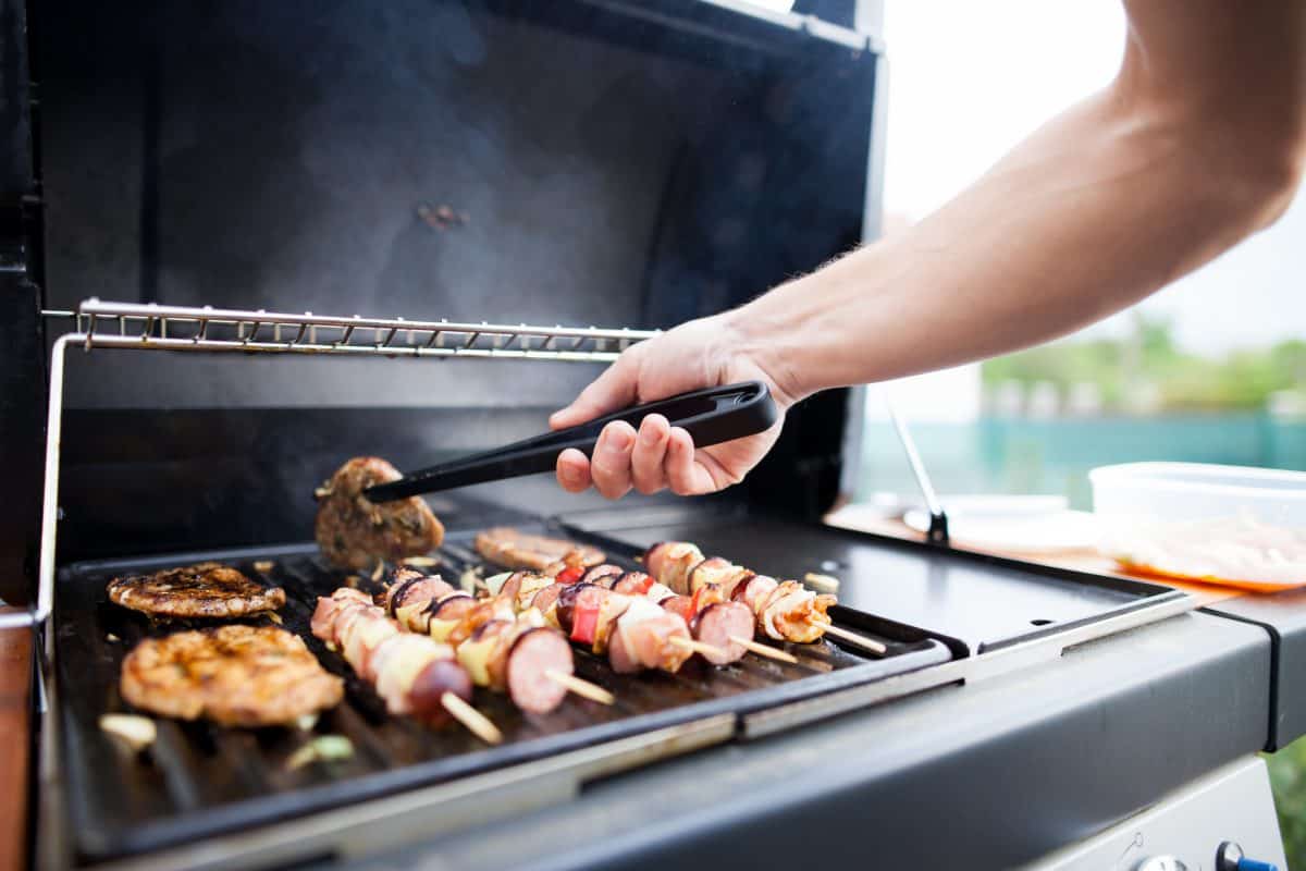 A man turning a burger on a gas grill that's grilling burgers and kebabs