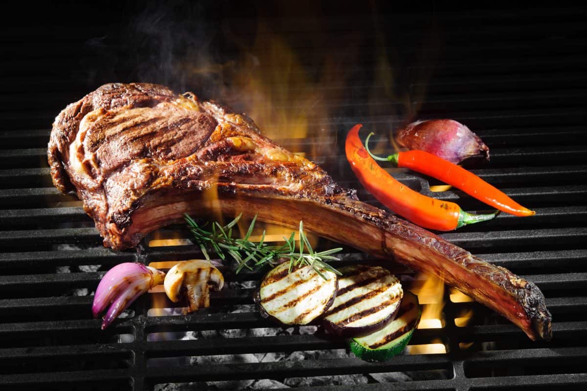 A tomahawk steak on a charcoal grill with some vegetable slices and a chili