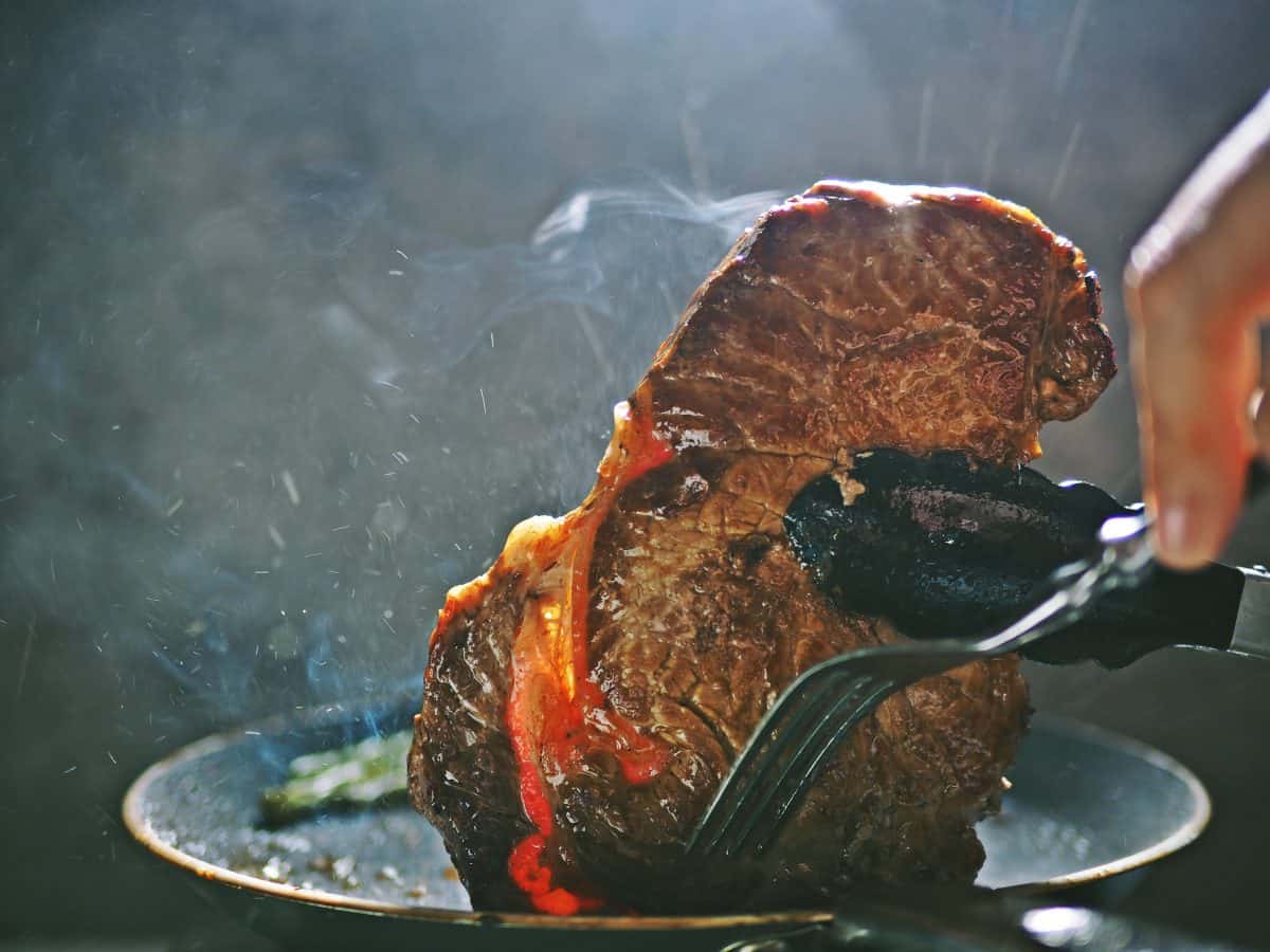 A steak being seared in a hot pan, being held on end by tongs